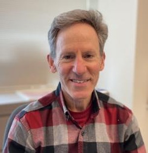 professor in a blue, white and, red button up plaid shirt in front of a white backdrop
